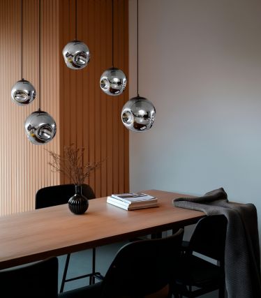 A stylishly furnished dining area with several Valentina pendant lights from Lindby. The spherical, chrome-plated lights hang at different heights above a light wood dining table. Black chairs, a vase with dried flowers, and subtle decorations create a modern, elegant ambience with warm lighting accents.