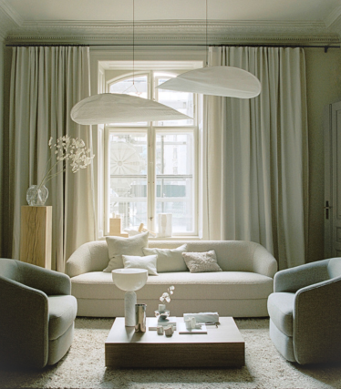 A serene, softly lit living room in neutral tones. A curved sofa and two armchairs in light beige surround a low wooden coffee table with minimalist decor. Above the seating area hang two large, sculptural white pendant lamps. Tall curtains frame a window that lets in gentle daylight, creating a calm and elegant atmosphere.