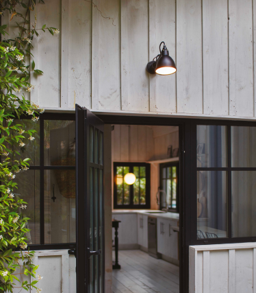 DCW outdoor wall light on a bright wooden facade with a view into a cosy interior.