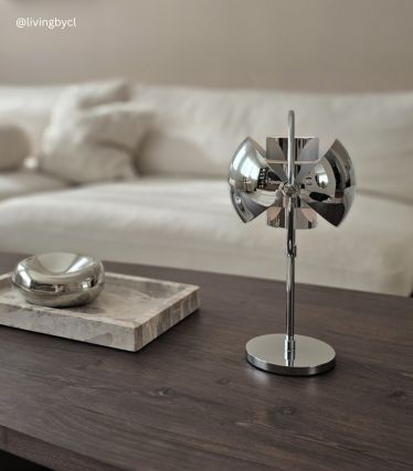 Chrome-plated designer table lamp on a wooden table in front of a light-coloured sofa.