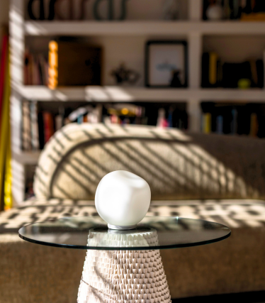 Small white table lamp on glass top, sunlight and bookshelf in the background.