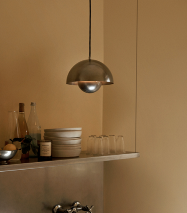 Pendant light made of polished metal above a shelf with ceramic bowls and glasses in front of a sand-coloured wall.