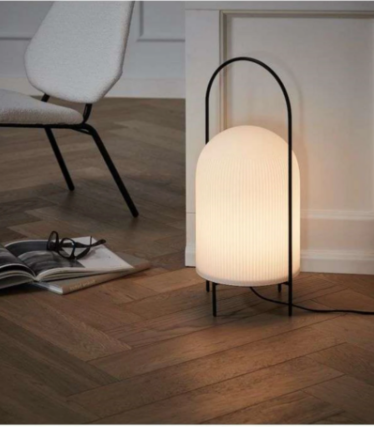 White ribbed lamp with a black handle frame on herringbone parquet next to an armchair.