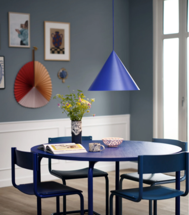 Blue cone-shaped pendant light above a round blue dining table with a bouquet of flowers, in a dining room with a blue wall and artwork.