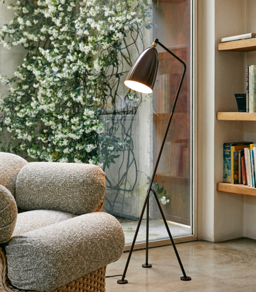 Slim GUBI floor lamp with dark lampshade lights up next to a structured armchair in a cosy room. A large window with flowering plants and a wooden shelf with books can be seen in the background.