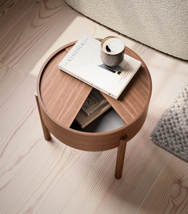 Round side table from Woud made of warm wood with an offset table top and open compartment. A Kinfolk book and a cup of coffee rest on the table on a light-coloured wooden base.