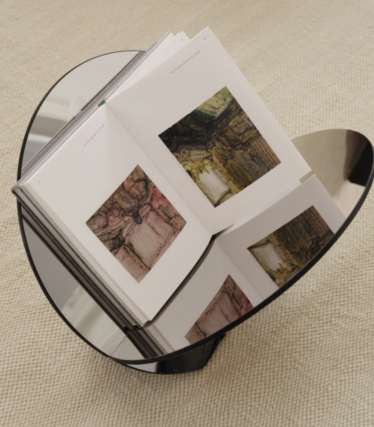 Round side table in dark glass with an open art book, on a beige rug.